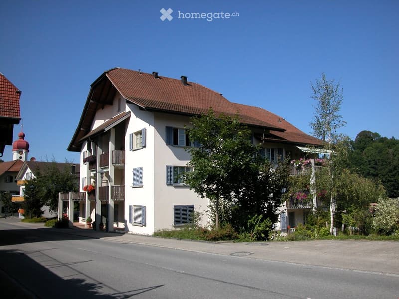 A two-story residential building with a red-tiled roof, white walls, multiple windows with shutters, and balconies adorned with flowers. The building is surrounded by greenery and trees, with a clear blue sky in the background.