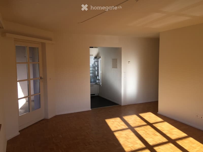 Empty room with wooden flooring and white walls, featuring a glass door and an open kitchen area with a refrigerator visible.