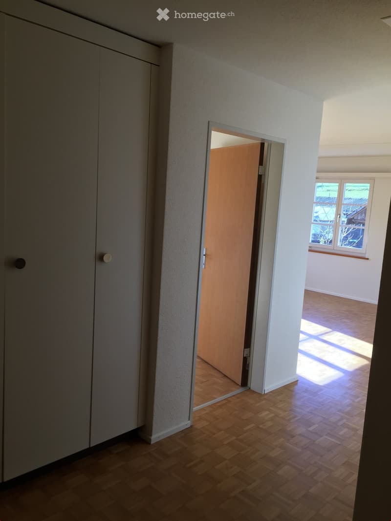 Empty hallway with wooden flooring, white walls, and a partially open wooden door leading to another room with a window showing outdoor view.