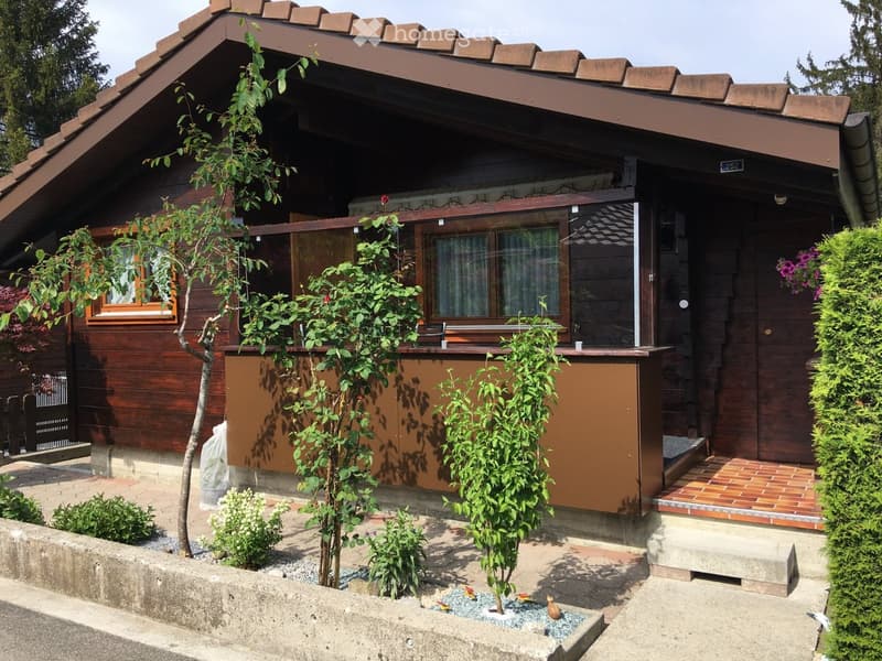 Two-story wooden house with a brown roof, large windows, and a small garden area with plants and a small decorative fountain.