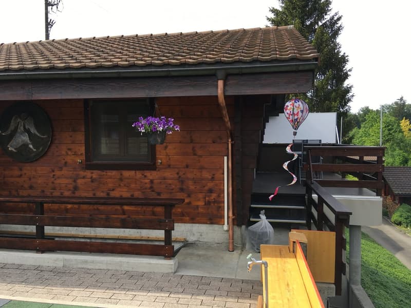 Wooden outdoor structure with a tiled roof, a window with a hanging flower pot, a staircase leading to a raised platform, and a small table and chairs near a water faucet.