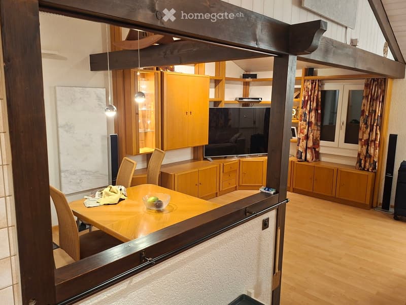 A well-lit living and dining area with wooden flooring, a dining table with chairs, cabinets, a TV screen, and curtains on the windows.