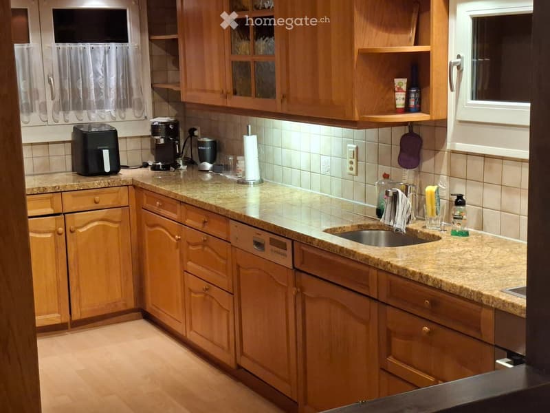A well-lit kitchen with wooden cabinets, granite countertops, and various appliances including a coffee maker, toaster, and dishwasher. A sink with a faucet is visible, along with a few bottles and cups on the counter.