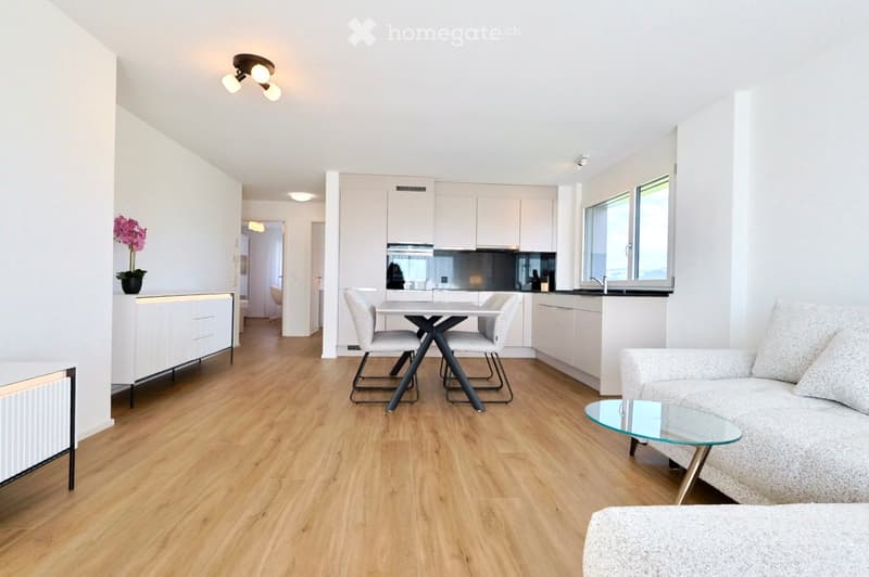 Modern, open-plan living and dining area with light wooden flooring, white walls, and contemporary furniture including a sofa, coffee table, dining table with chairs, and kitchen cabinets.