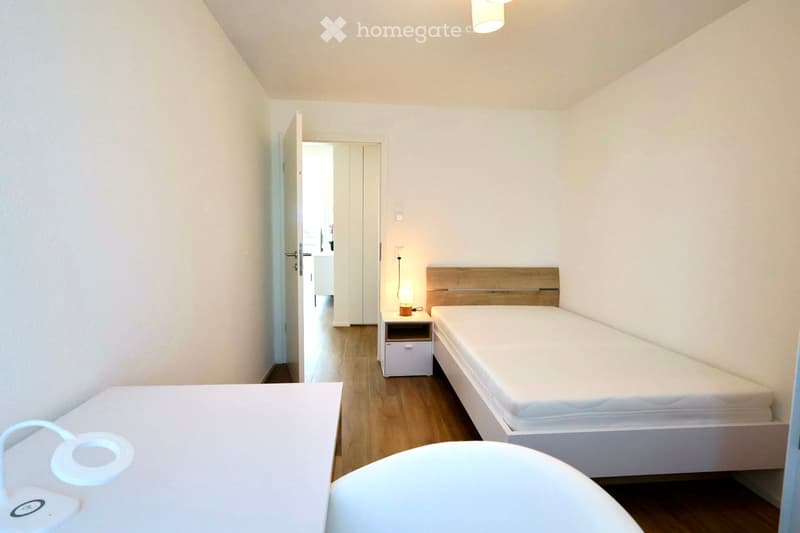 A small, modern bedroom with a single bed, a nightstand with a lamp, and a drawer unit. The room features light wooden flooring and white walls.