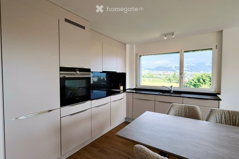 Modern kitchen with white cabinets, black countertops, and a large window offering a scenic view. Includes a dining table with chairs, an oven, and a sink with a faucet.
