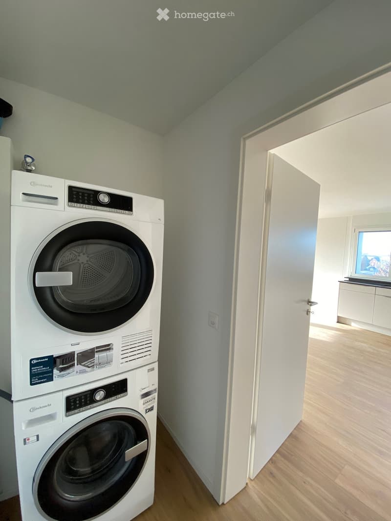 A small, well-lit room with light wooden flooring and white walls, featuring a stacked washing machine and dryer unit.