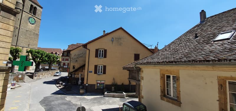 A picturesque European town square with a tall clock tower, traditional stone buildings, a restaurant with outdoor seating, and a clear blue sky.