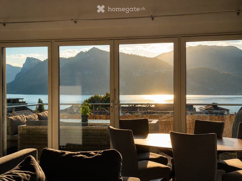 A modern living space with large windows offering a scenic mountain and lake view. The room features a sofa, a dining table with chairs, and a small potted plant on the balcony.