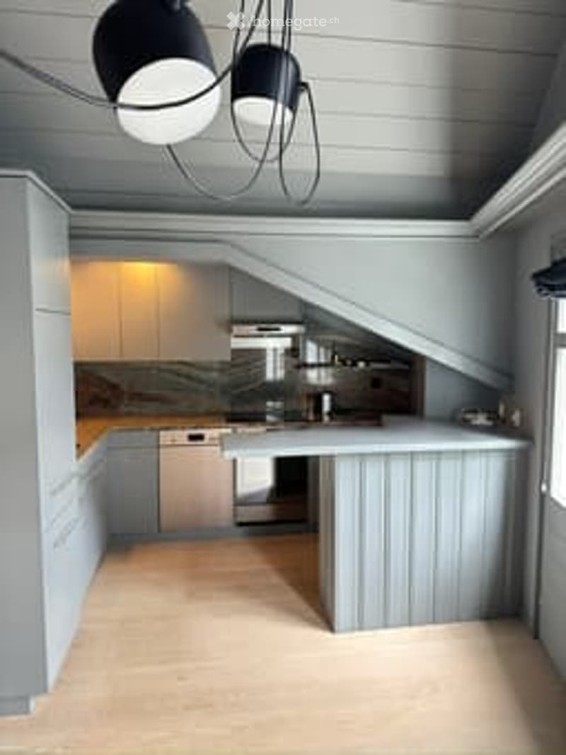 Modern kitchen with light grey cabinetry, a central island, and stainless steel appliances including a stove and dishwasher. The space features a sleek, minimalist design with a hanging pendant light fixture above the island.