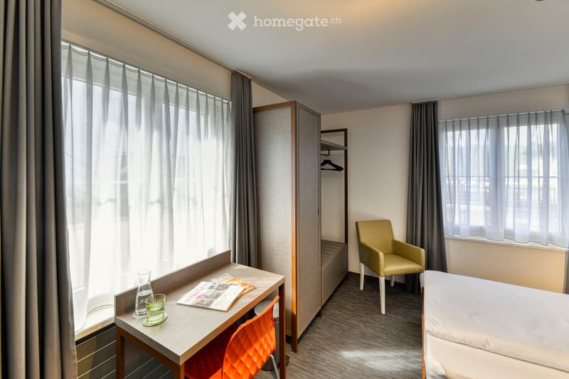 A modern, furnished room with a bed, a desk, a chair, and a wardrobe. The room features large windows with curtains, a grey carpeted floor, and a contemporary design.