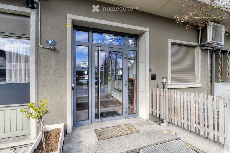 Entrance to a building with a glass door labeled "Hotel Casan," a small planter with a plant, and a wooden fence on the right side.