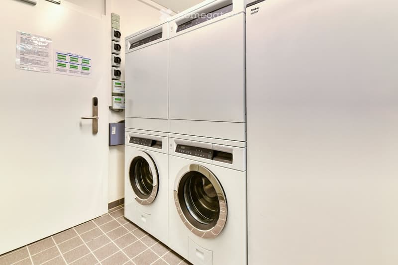 A clean, modern laundry room with white tiled flooring and white walls. Two stacked washing machines and dryers are positioned side by side, with a control panel above each unit. A door with a silver handle is visible on the left side.