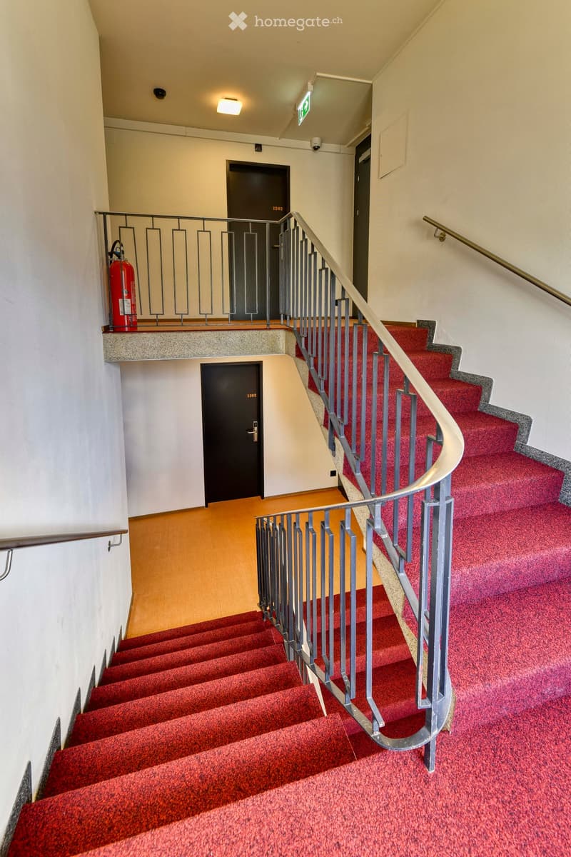 Staircase with red carpeted steps, metal railings, and doors labeled 1102 and 1202, with a fire extinguisher on the landing.