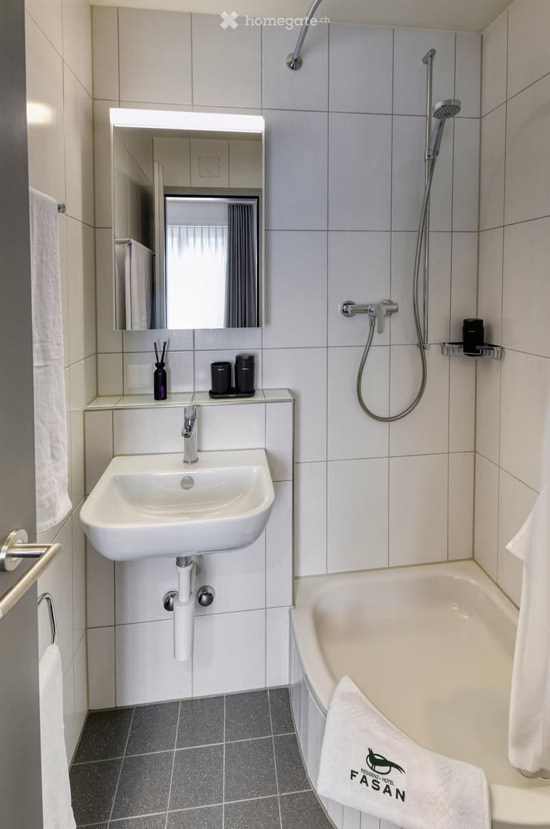 Modern bathroom with a white sink, mirror, bathtub, and showerhead. Includes a towel on the edge of the tub and a small shelf with toiletries.