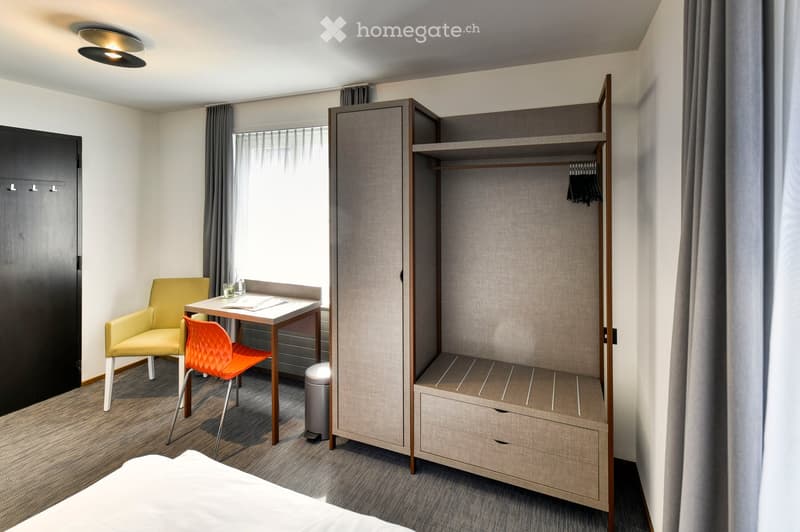 A modern, compact bedroom with a bed, a wardrobe, a small table with chairs, and a stool, all set in a cozy, well-lit space.