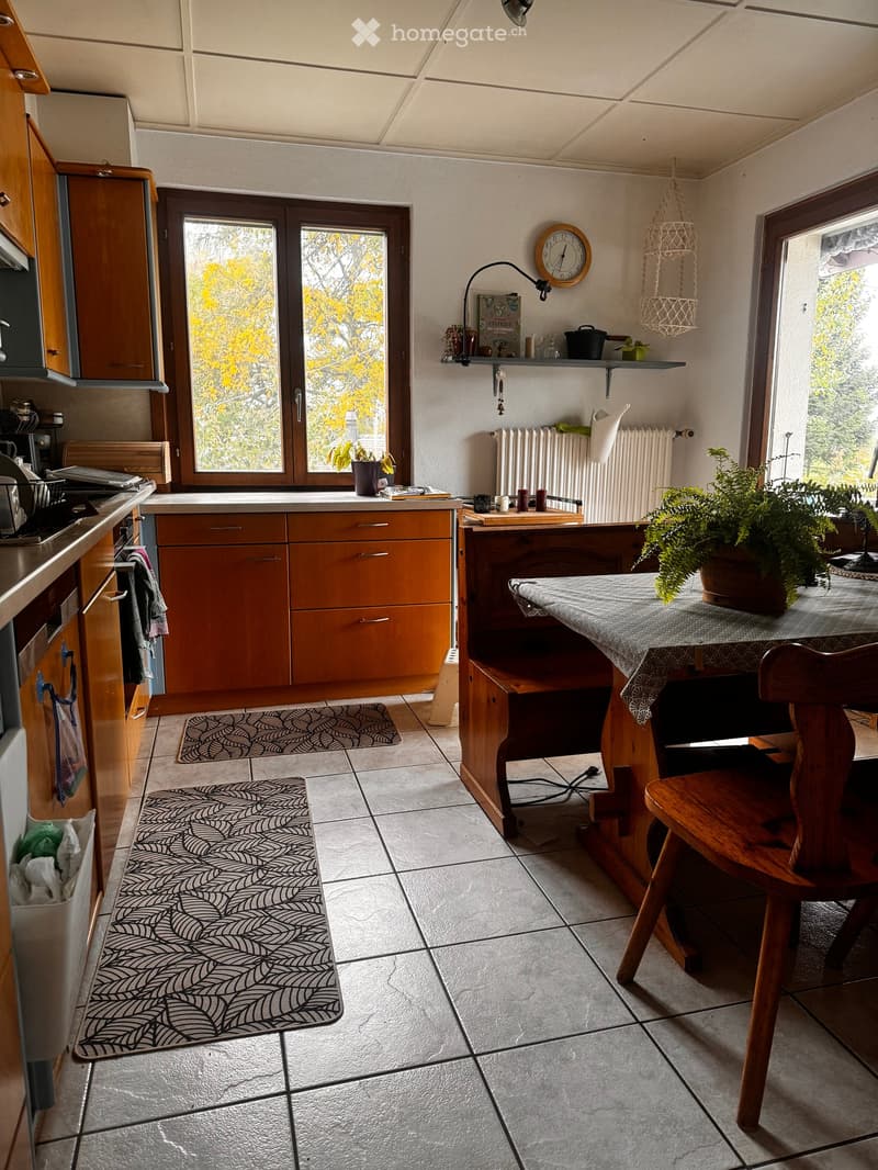 The kitchen features wooden cabinets, a tiled floor, and a dining area with a table and chairs. There is a radiator, a clock, and various kitchen appliances visible.