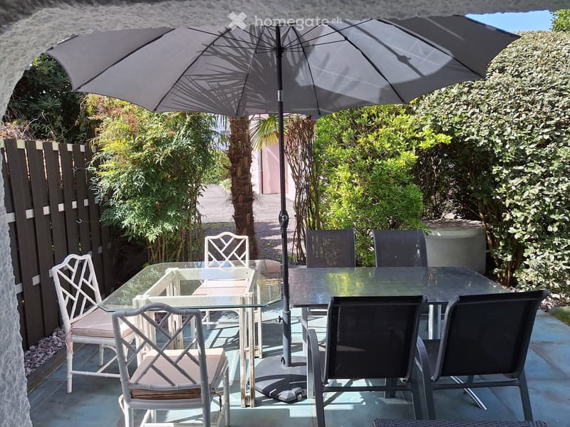 Outdoor patio with glass table, white and black chairs, large umbrella, surrounded by greenery and a stone wall.