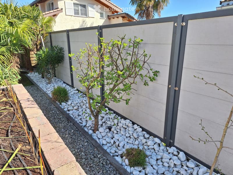 Modern garden area with a small tree, shrubs, white pebbles, and a low concrete wall, bordered by a grey privacy fence.