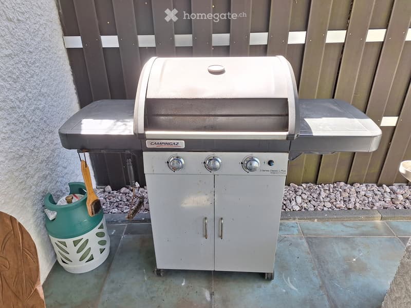 A modern gas grill with control knobs and a storage cabinet, placed on a tiled patio next to a gas tank and a brush.