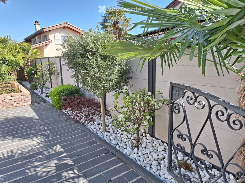 A well-maintained garden area with a paved walkway, decorative white pebbles, small trees, shrubs, and a wrought-iron railing leading to a house entrance.