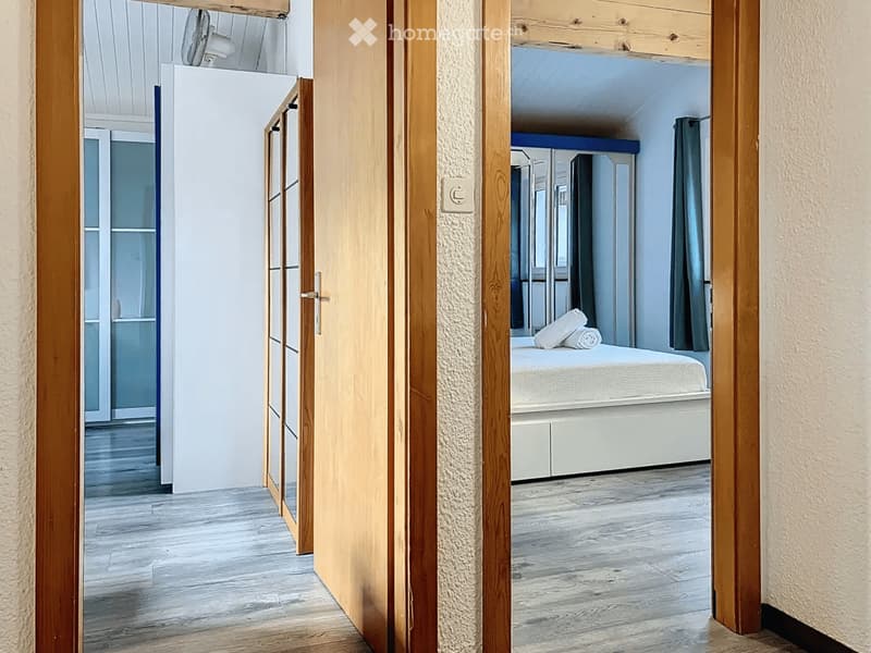 Modern bedroom with light wooden flooring, white walls, and a neatly made bed with pillows. The room features a large window with dark curtains and a built-in wardrobe with mirrored doors.