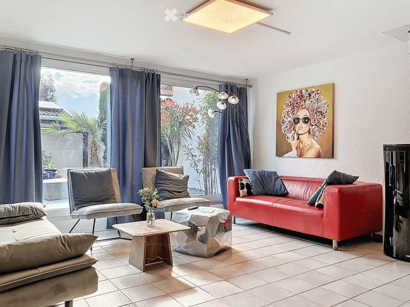 Modern living room with large windows offering a scenic view, featuring a red leather sofa, a grey armchair, a coffee table, and a floor lamp.