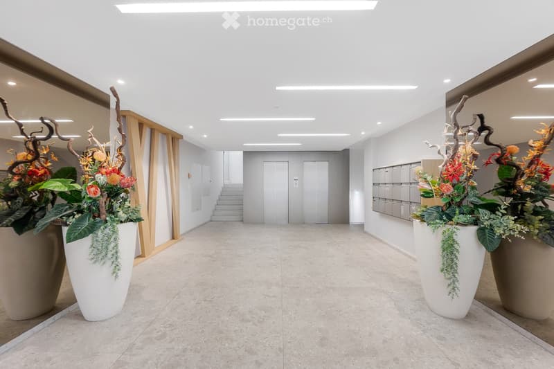 Modern hallway with white walls, grey tiled floor, and large floral arrangements in white pots on either side. Features a staircase leading upwards and elevator doors at the end.