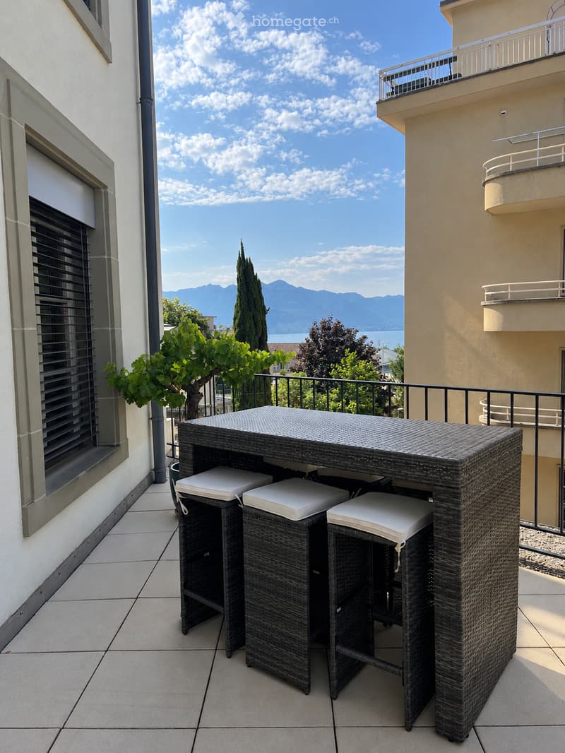 Balcony with a wicker table and three stools, overlooking a scenic view of mountains and a lake.