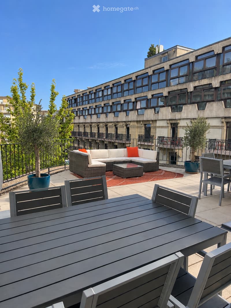 Modern rooftop terrace with a grey sectional sofa, orange cushions, a coffee table, and outdoor dining set including a table and chairs. Potted trees and a cityscape view are visible in the background.