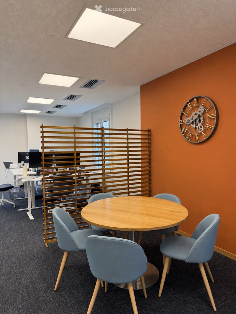 Modern office space with a round wooden table surrounded by blue chairs, a wooden screen divider, a computer screen, and a clock on the wall.