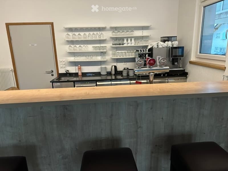 Modern bar area with wooden counter, shelves holding glasses, coffee machine, and stools.