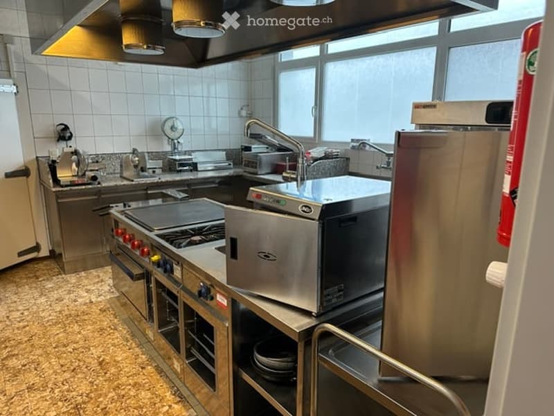 Commercial kitchen with stainless steel appliances, including a stove, fryer, refrigerator, sink, and microwave. Large windows provide natural light.