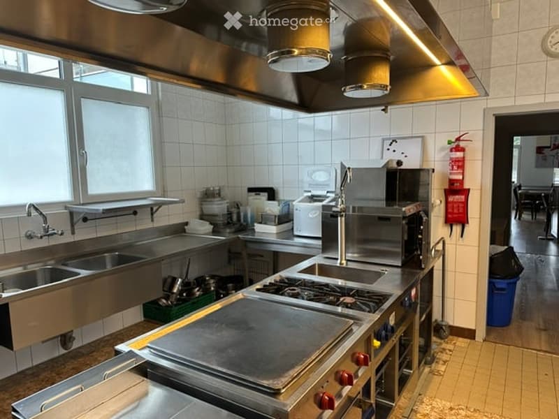 Commercial kitchen with stainless steel appliances, including a sink, microwave, oven, stove, and refrigerator. The space features large windows, tiled walls, and a well-organized counter area.
