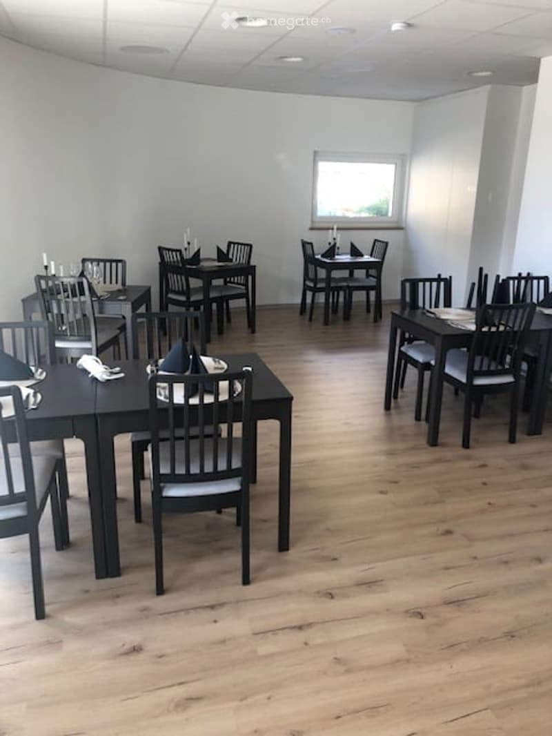 A modern, minimalist restaurant or dining area with wooden floors, black tables, and matching chairs arranged neatly. Each table is set with napkins, candles, and glasses, creating a clean and inviting atmosphere.
