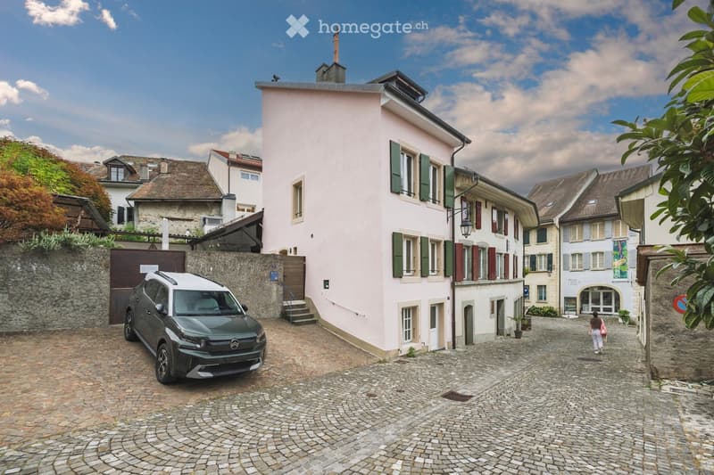 A charming pink building with green shutters and a stone staircase leading to the entrance, situated in a narrow cobblestone street with a parked car and surrounding historic architecture.
