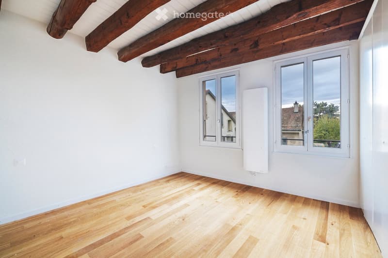 Empty room with wooden flooring, white walls, and exposed wooden beams on the ceiling. Two large windows with white frames offer natural light and views of the outside.