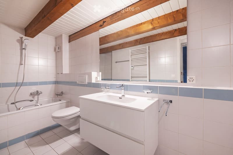 Modern bathroom with white tiles, a large sink, toilet, bathtub, and a showerhead. Features exposed wooden beams on the ceiling and a large mirror above the sink.
