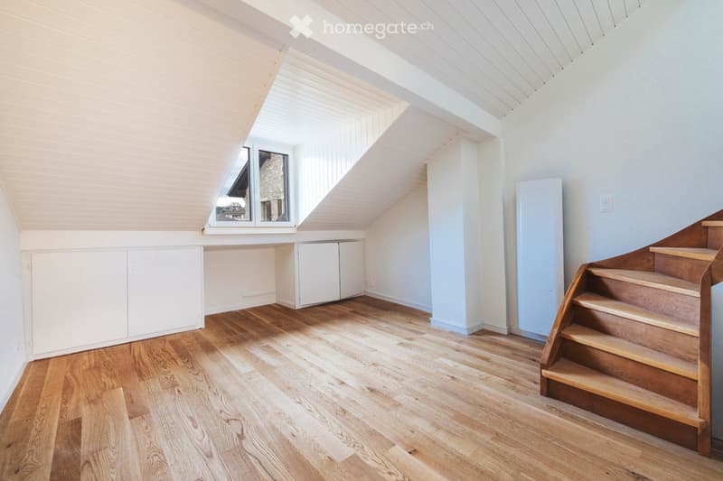 Bright, airy attic space with wooden flooring, white walls, built-in cabinets, and a wooden staircase leading upwards. Large windows allow natural light to fill the room.