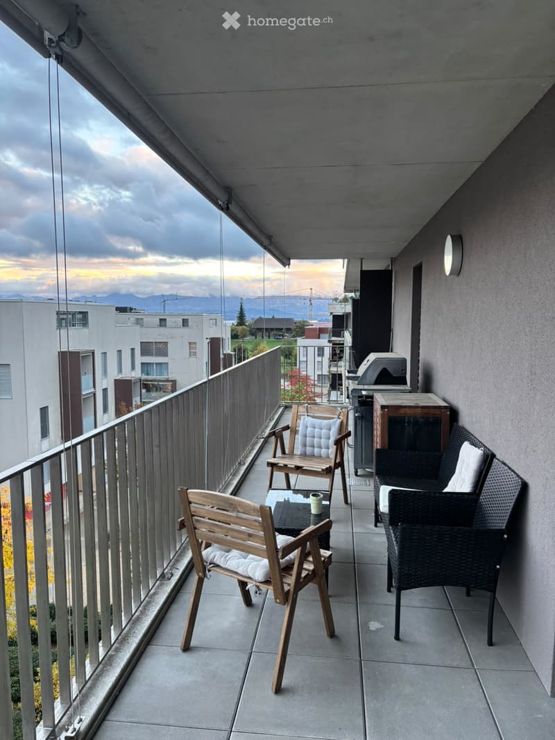 Balcony with wooden chairs, a wicker chair, a small table, and a cup, overlooking a cityscape with mountains in the background.
