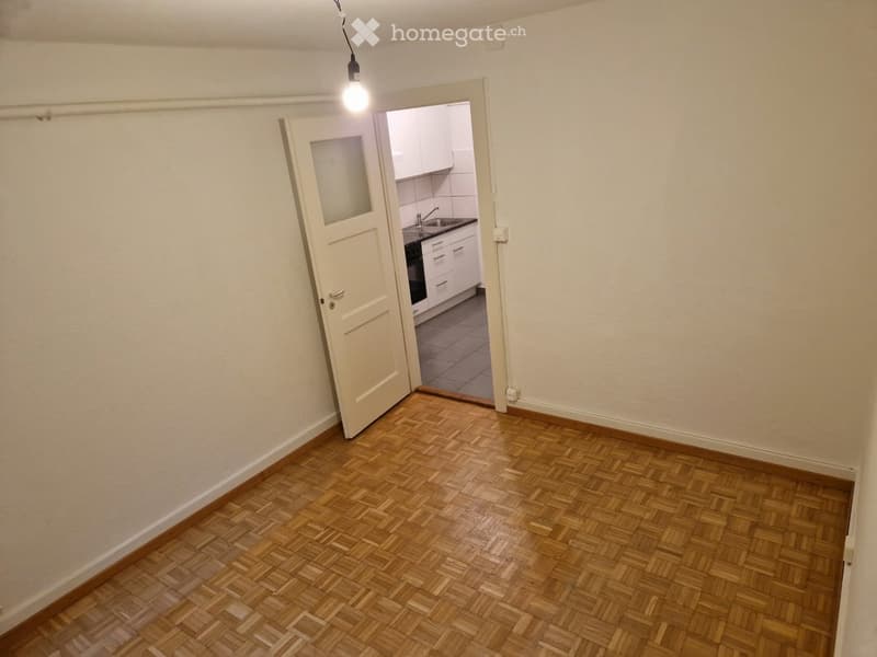 Empty room with wooden herringbone flooring, white walls, and an open door leading to a small kitchen area.