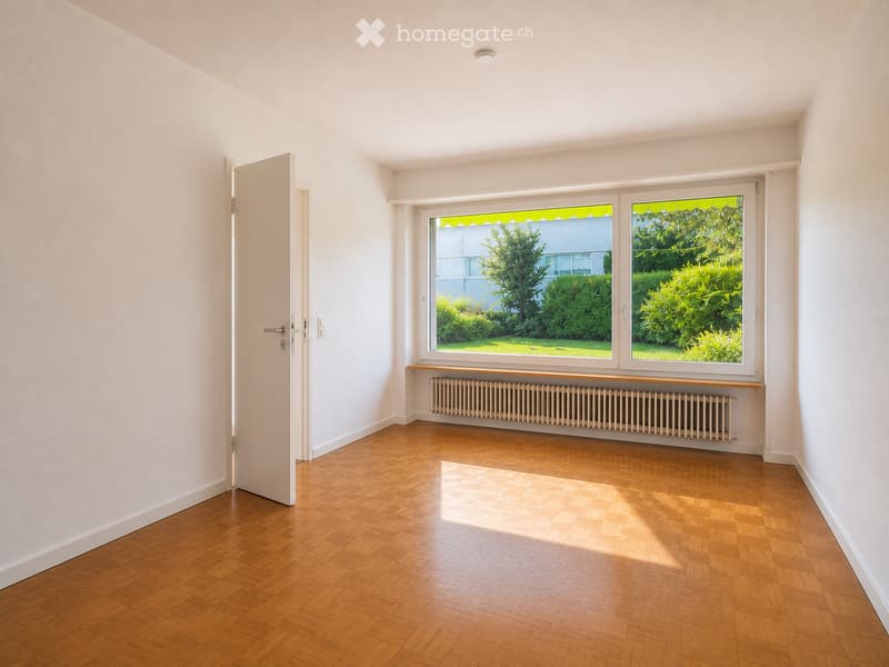 Empty room with wooden flooring, white walls, large windows with green outdoor view, radiator, and an open door leading to another space.