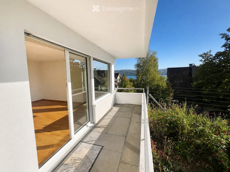 Modern balcony with large glass sliding doors, wooden floor inside, tiled balcony floor, metal railing, and scenic view of a lake and mountains.