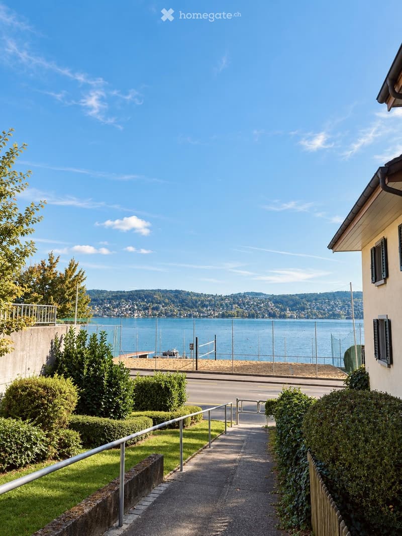 Pathway leading to a waterfront view with a clear blue sky, distant hills, and a fenced beach area.