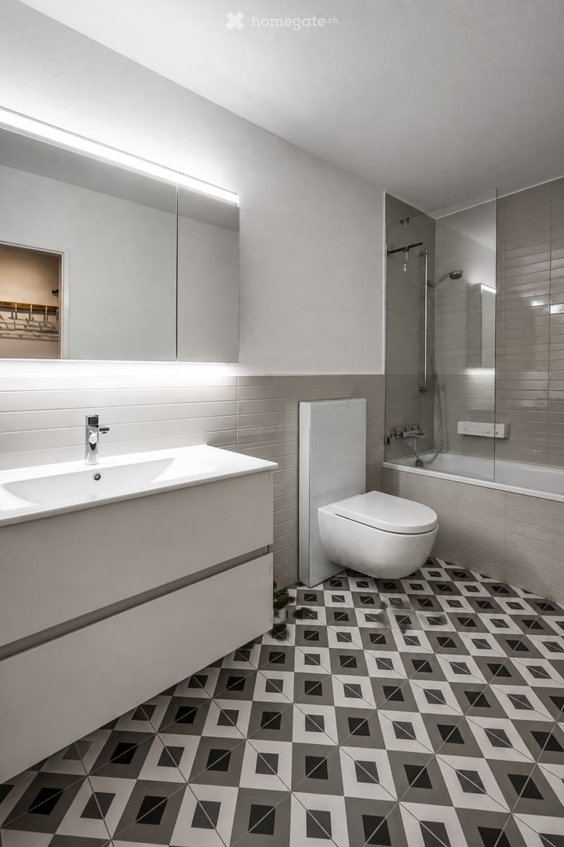 Modern bathroom with white tiles, geometric patterned floor, sink, toilet, bathtub, and mirror.