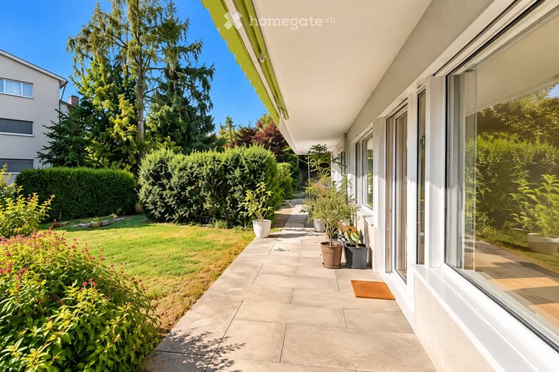 A well-maintained garden with a paved pathway, potted plants, and a neatly trimmed lawn, surrounded by trees and shrubs.