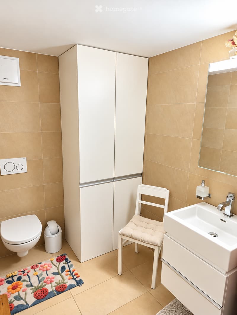 Modern bathroom with beige tiles, white toilet, sink, mirror, and a small white chair with a cushion. Features a floral rug and a white cabinet.
