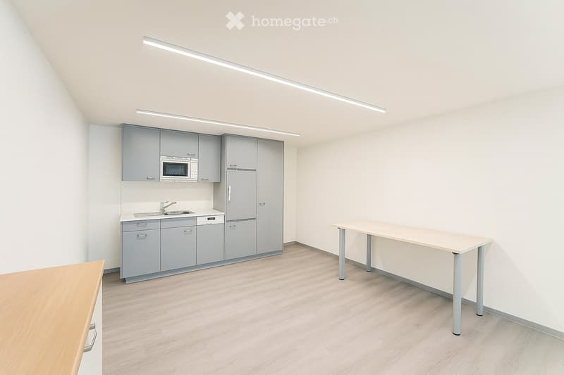 Modern, minimalist kitchen with grey cabinets, a refrigerator, microwave, sink, and a wooden table.