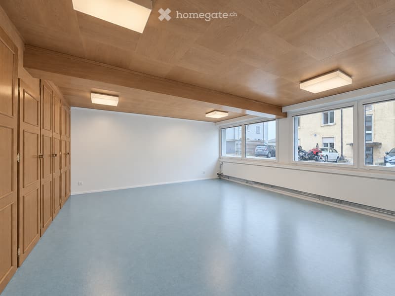Empty room with light blue carpeted floor, wooden paneled ceiling, white walls, large windows, and a wooden door.