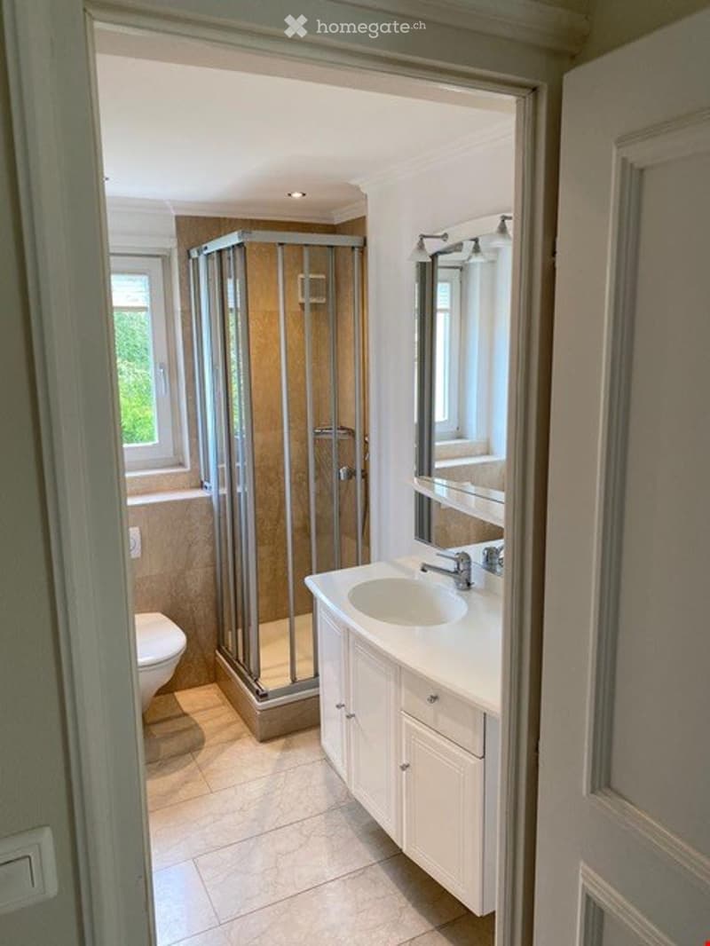 Modern bathroom with a white sink, mirror, and a glass-enclosed shower stall. The space features light-colored tiles and a window providing natural light.